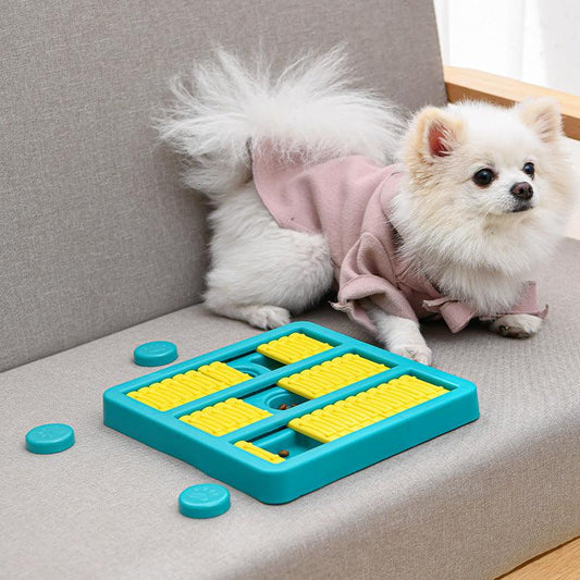A small dog wearing a pink shirt sitting next to a blue and yellow interactive food dispensing dog toy with caps on the couch.-  The PetRiot